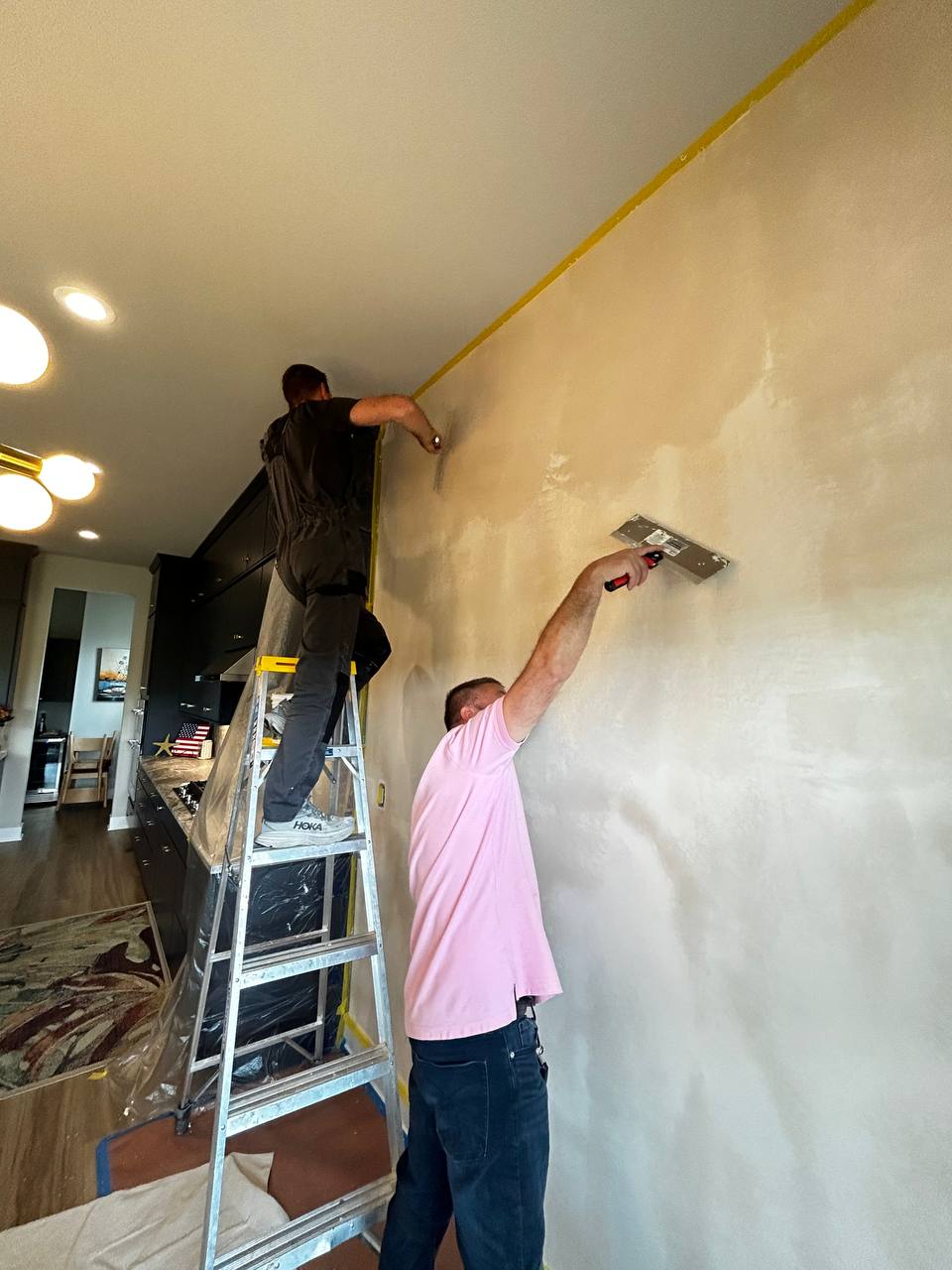 Professional installers on ladders applying smooth white wallcovering to an office wall, using laser lines for precise alignment.
