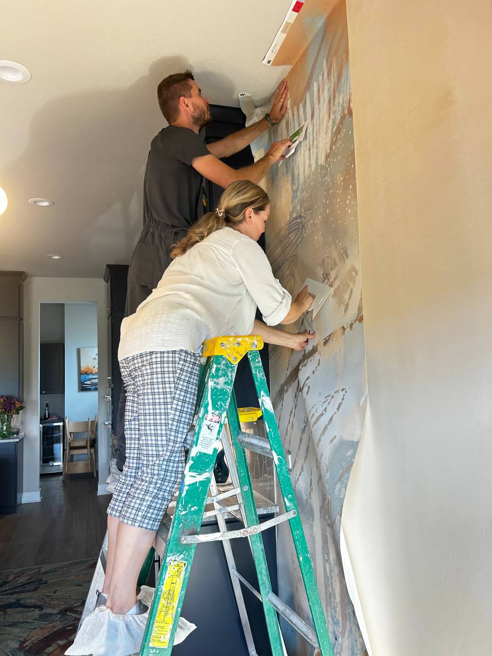 Installers applying a large-scale blue and cream checkered (gingham) pattern wallpaper to an accent wall near the ceiling trim and window.
